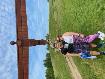 A young man and woman stand in front of Angel of the North sculpture smiling to camera