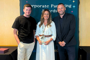 A woman holds a glass trophy standing between two men, all three smiling to camera