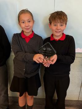 A girl and boy wearing grey, black and red school uniform, smiling and holding a glass award.