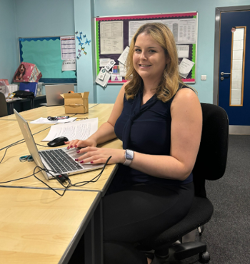 A woman sits at a desk using a laptop smiling to camera