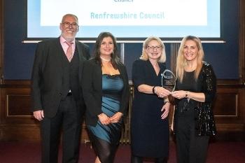 A man in a suit stands alongside three women in evening wear all smiling to camera. The two women on the right are both holding a glass trophy.