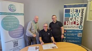 Three men, one sitting at a table with a document and the other two standing at each side looking to camera