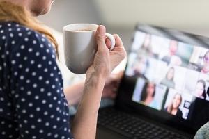 A woman holding a cup of tea sits in front of a laptop which displays an online team meeting with several attendees