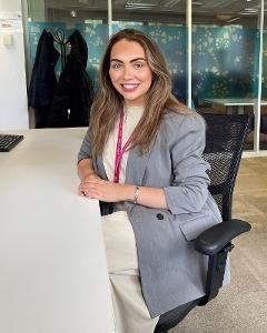 A woman with long dark hair sits on a chair by a desk smiling to camera